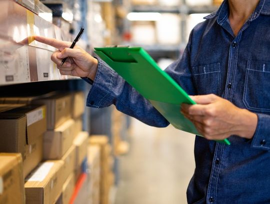 A person in a denim shirt holding a green clipboard and examining various boxes that are stacked on warehouse shelves.