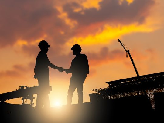 Two construction workers standing on a construction site, shaking hands with the sunlight casting a shadow over them.