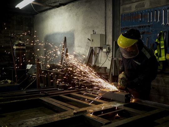 A worker wearing a face shield and gloves grinding a metal frame as bright sparks scatter across a dark workshop.