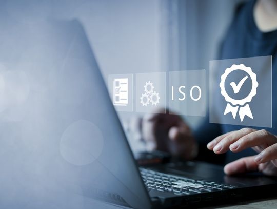 A close-up of a woman using a laptop with holographic icons for a checkmark badge and "ISO" floating in front of her.