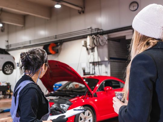 A woman in work clothes writing on a clipboard beside another woman near a red car in an auto repair shop.