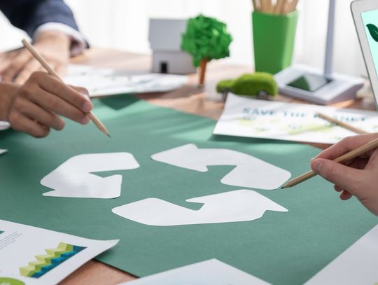 Hands sketch recycling icon during eco planning meeting, with charts, reports, and laptop displaying “Think Green” message.