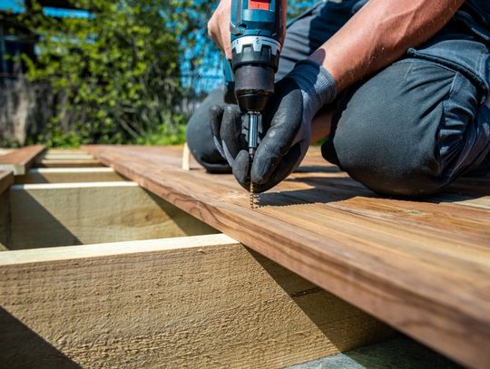 A close-up of a person using an electric drill to fasten a screw into wooden deck boards over exposed framing.