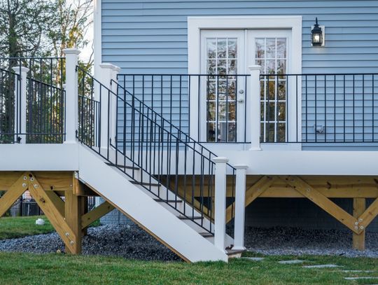 A raised deck attached to the back of a home. The deck has stairs with white posts and metal railings leading to the ground.