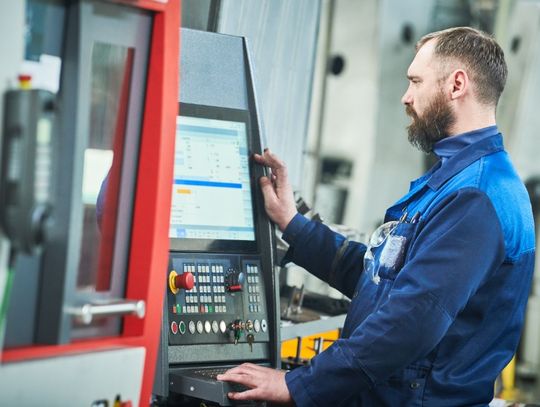 A man stands in front of the control panel and screen of a CNC machine. He looks through various settings.