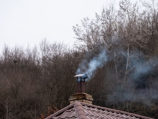 A cloud of smoke billows out from a brick chimney on a metal tiled roof on top of a residential building.
