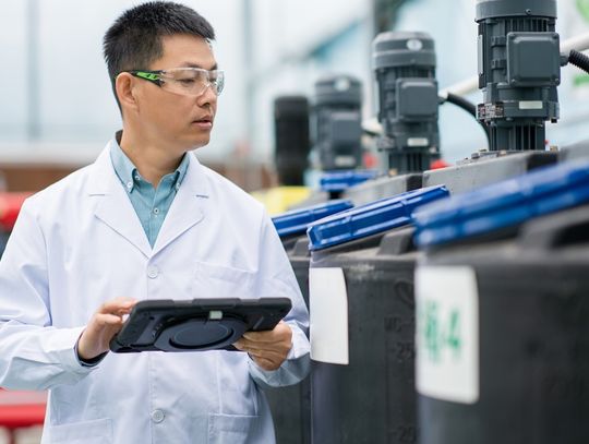 A man in a long white coat and goggles is observing black and blue containers. He’s holding a tablet in his hand.