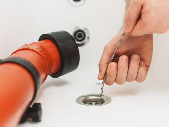 A close-up view shows a person placing a metal drain snake down the drain of a white bathtub. An orange tube is in the tub.