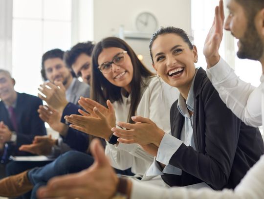 Several people in business attire sitting in a row, clapping and smiling at the person on the end, who has his hands outstretched.