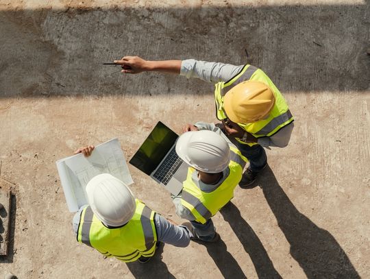 Three construction workers are standing next to each other. One is holding a laptop while another is pointing.