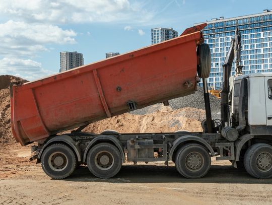 A white dump truck with a raised orange bed standing on dirt beside piles of soil and gravel with city buildings behind it.
