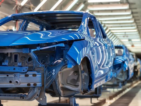 Blue unfinished car bodies move along a brightly lit automotive factory assembly line during vehicle production.