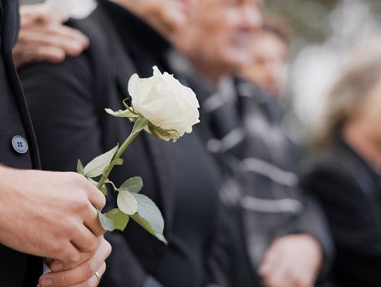 A group of people wearing black mourning outfits holds a white rose while attending a memorial service.
