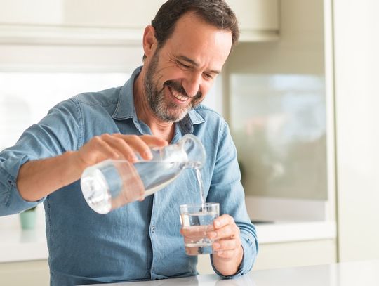 A man smiles as he stands in his kitchen and pours water from a bottle into a clear glass. He wears a denim shirt.