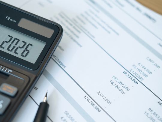 A calculator has the number "2026" on its screen as it sits on a stack of bills. A pen sits next to it.