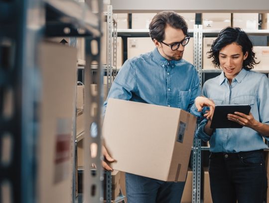 Two employees checking stock levels on a tablet. One employee holds a cardboard box and the other has the tablet.