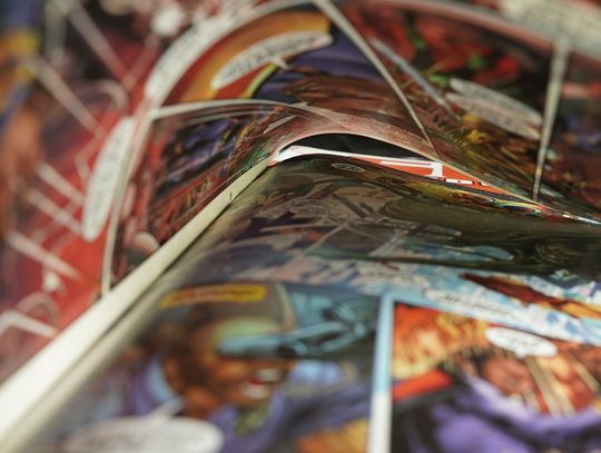 An open comic book curls across a table, showing colorful superhero panels in a soft, close-up blur.