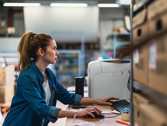 A woman working on her computer as a desk. She is surrounded by tons of boxes on shelves and on the floor.