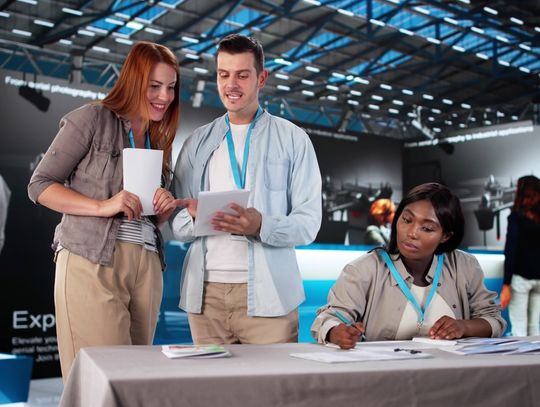 A group of three people wearing light blue lanyards and holding flyers prepare to present in a trade show conference.