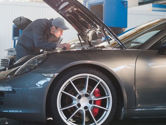Male mechanic in a gray uniform is inspecting the hood of a gray Porsche sports car in an auto garage shop.