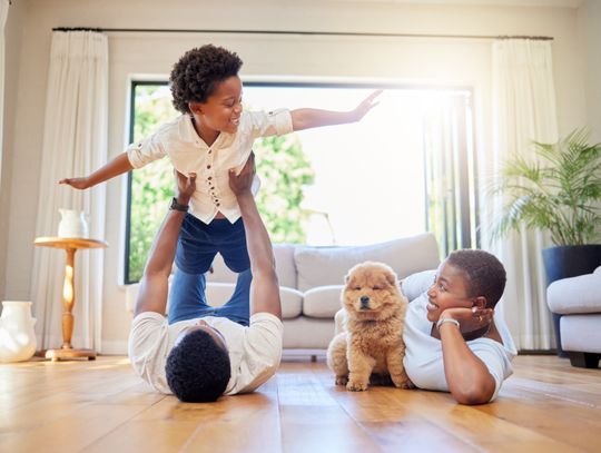 A dad and mother lying on a living room hardwood floor with their child raised on legs and a small dog beside them.