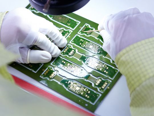 A point-of-view close-up of a person with gloves working on a green flexible electronic circuit board.