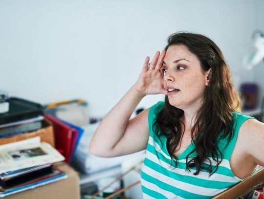 A woman wearing a green and white shirt holds her head while looking at piles of clutter. She's also holding a box of items.