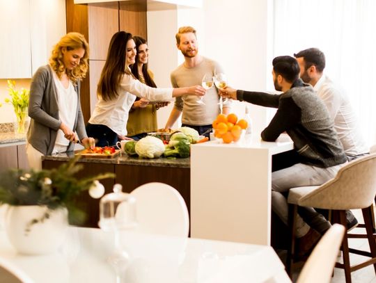 Four people raising glasses of white wine over a kitchen table with plates of food and blurred lights behind them.