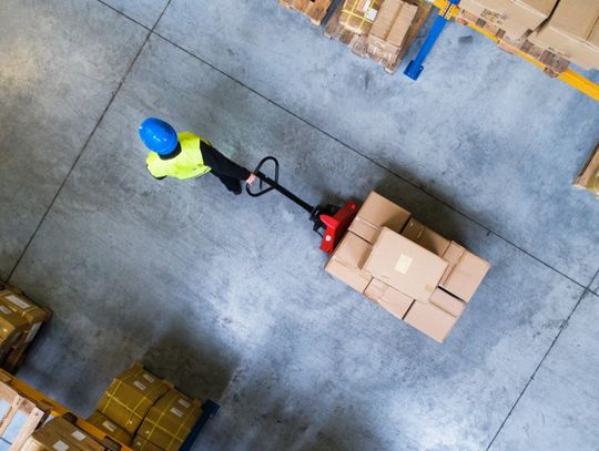 An aerial view of a warehouse employee in PPE gear rolling a dolly behind them. The dolly has boxes on it.