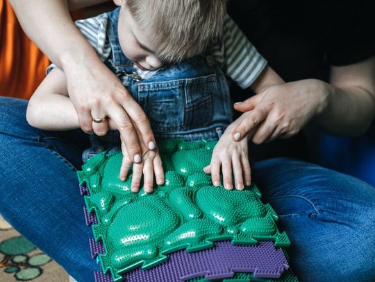 A young boy sits in the lap of a woman while being guided to participate in an activity with a sensory mat.
