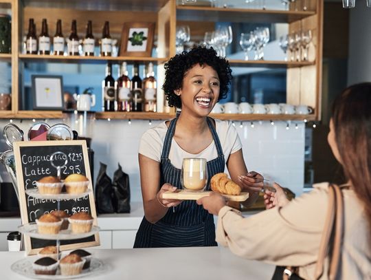 A smiling female barista hands a latte and pastry to a female customer at a cozy coffee shop counter.