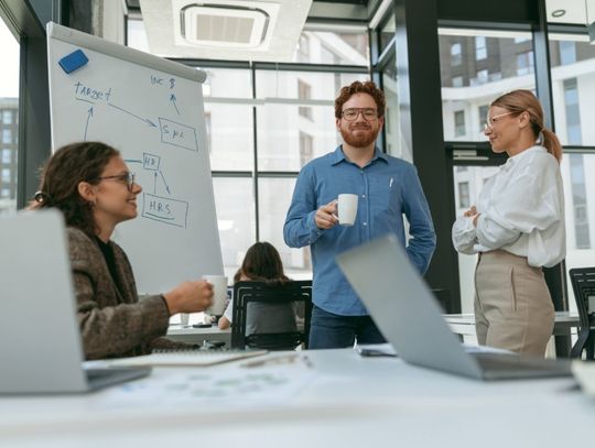 Coworkers gather for a casual office discussion beside a flip chart, while laptops sit open in the foreground.