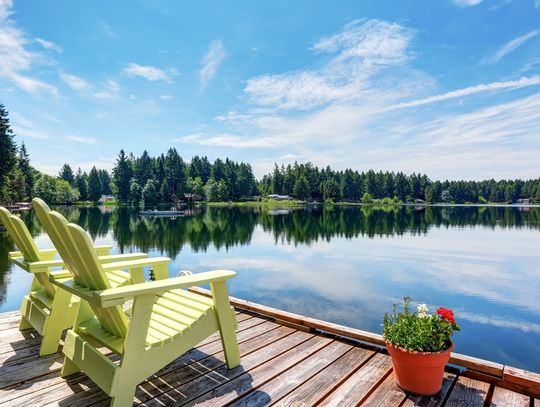 A wooden pier with two yellow Adirondack chairs and a potted plant, overlooking a lake surrounded by trees.