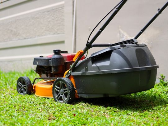 An orange and black push lawn mower with a rear grass collection bag resting on green grass beside a concrete wall.