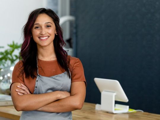 A smiling woman wearing an orange shirt and a gray apron stands in front of a reception area with crossed arms.