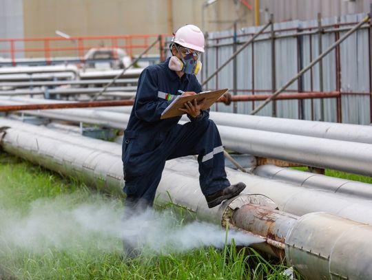 A male worker in full PPE writes on a clipboard and stands with his foot on a gas line that is emitting a stream of steam.