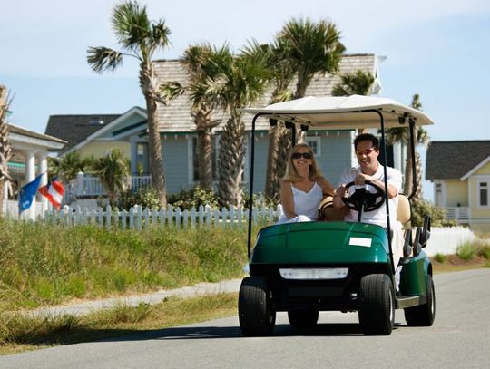 Two people in a golf cart on the road. There are houses, fences, and palm trees in the distance.