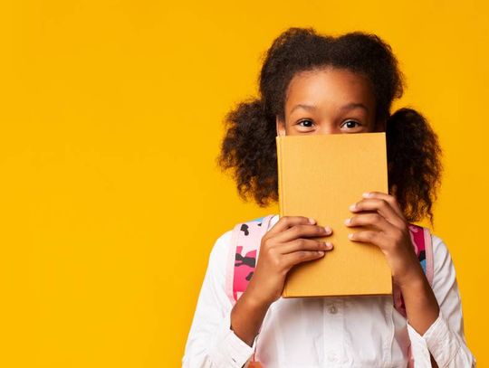 A young girl holds a yellow book in front of the lower half of her face in front of a yellow background.