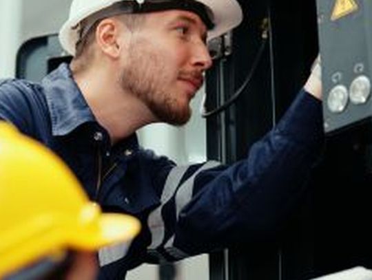 Two male production engineers in safety wear are assisting with the adjustment and maintenance of a machine in the facility.