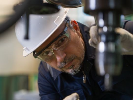 A close-up of a man in a hard hat and safety goggles with gloves crouching to inspect an industrial drill machine.