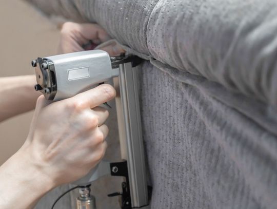 A person's hands holding a metal upholstery stapler against gray sofa fabric with a blurred fabric edge behind it.