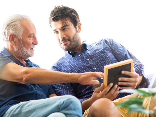 Two men seated on a couch look at a tablet together while the older man reaches toward its screen.