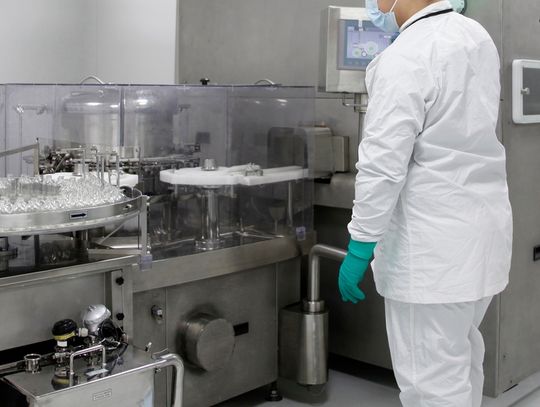 A rear view of a person in a lab coat, protective gloves, and a face mask standing in front of a drug production line.