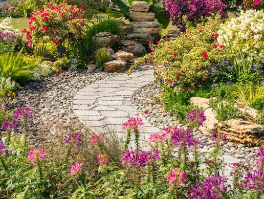 A stone path leading through a residential garden. The path is surrounded by vibrant grass and colorful flowers.