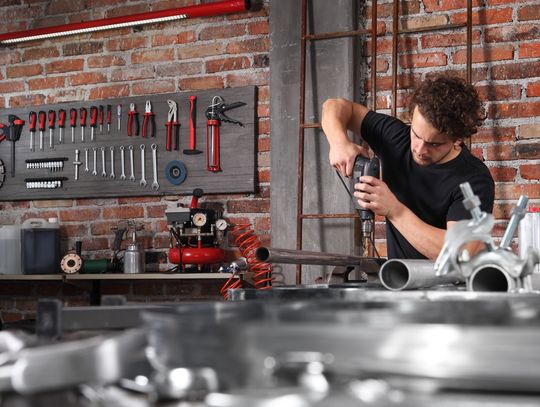 A man using a drill on a metal tube in his workshop. There is a lot of metal lying around, and some tools hung up on the wall.