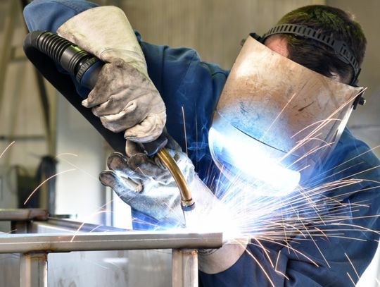 A person wearing a welding helmet, blue work jacket, and protective gloves welding a metal structure as sparks fly.
