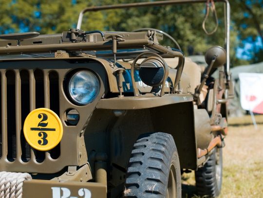 Half of the front part of an olive green jeep with a yellow badge on the grill. The jeep sits on grass near some tents.