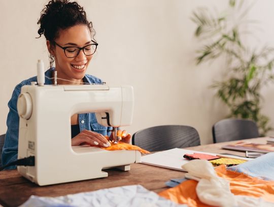 A young woman using a sewing machine and smiling at her table. The table is covered in bits of other fabric.