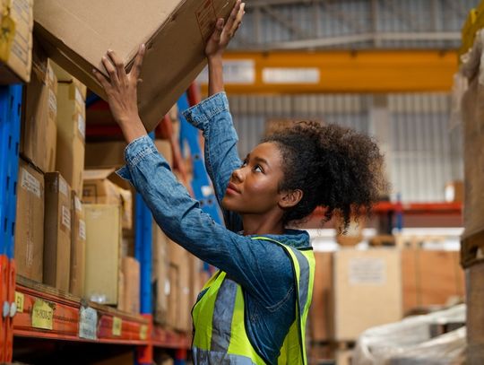 A warehouse worker wearing a safety vest reaches up to place a box on a high shelf among stacked packages.