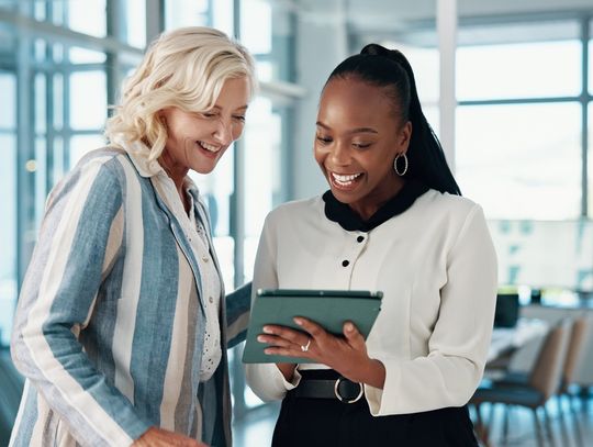 Two happy women business partners are looking at a tablet happily and pointing at the screen outside a conference room.